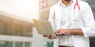 Arzt mit einem Stetoskop um den Hals vor einem Krankenhaus. Er hält einen aufgeklappten Laptop in den Händen.