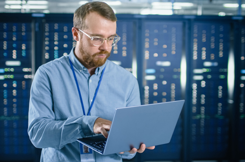 Mitarbeiter steht in Serverraum mit aufgeklapptem Laptop vor sich.
